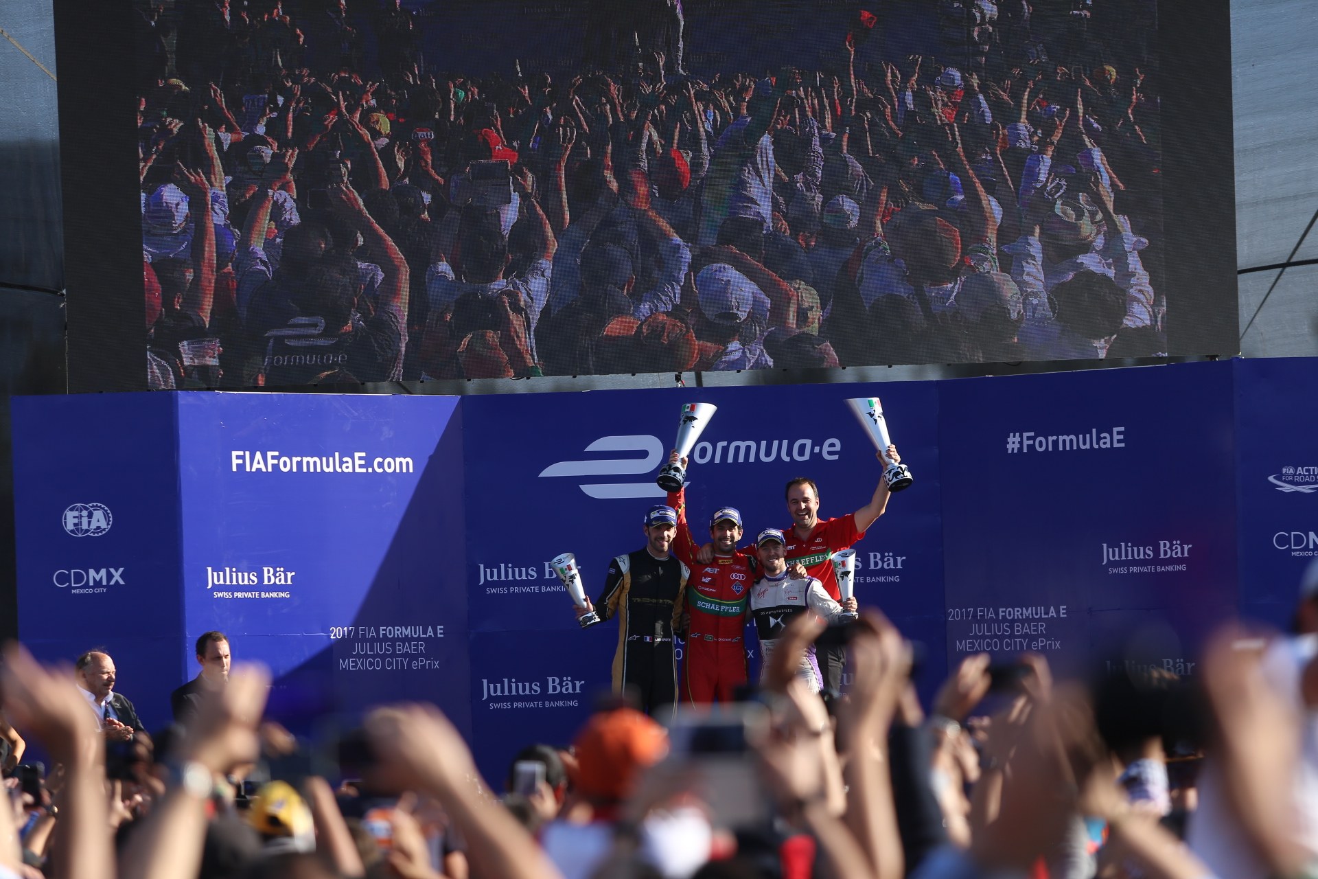 Gruppe von Menschen auf einem Formel-E-Podium in Mexiko-Stadt mit Tassen, Sponsor-Plakaten und einem Hintergrundbild.
