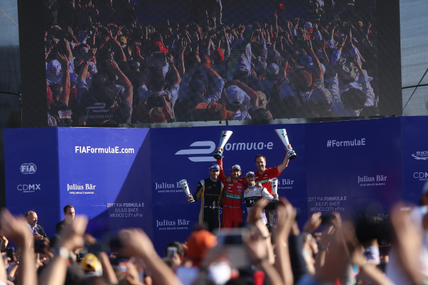 Gruppe von Menschen auf einem Formel-E-Podium in Mexiko-Stadt mit Tassen, Sponsor-Plakaten und einem Hintergrundbild.
