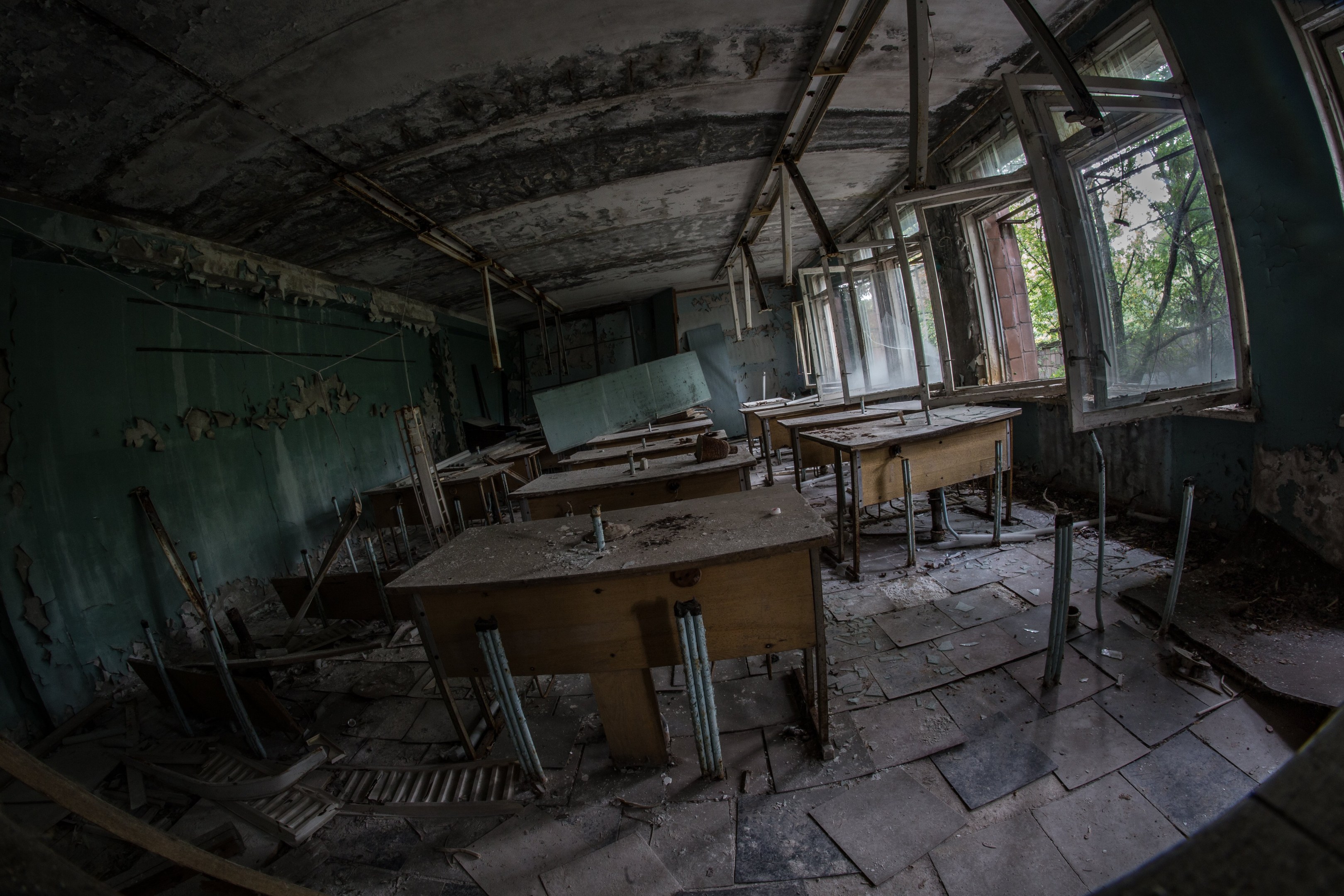 Verlassener Klassenraum in Pripyat, Tschernobyl, Ukraine mit Tischen, Stühlen, abblätternder Wandfarbe, kaputten Fenstern und sichtbarer Decke und Bäumen draußen.