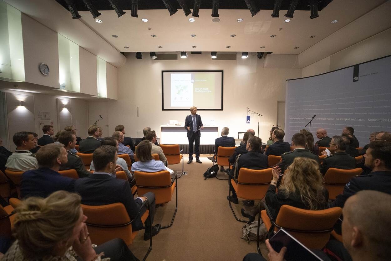 A man standing with a microphone addresses a seated audience in front of a projector screen displaying "Koninklijke Landmänch - The Future of the Future," with a clock, lights, and ceiling fixtures visible.