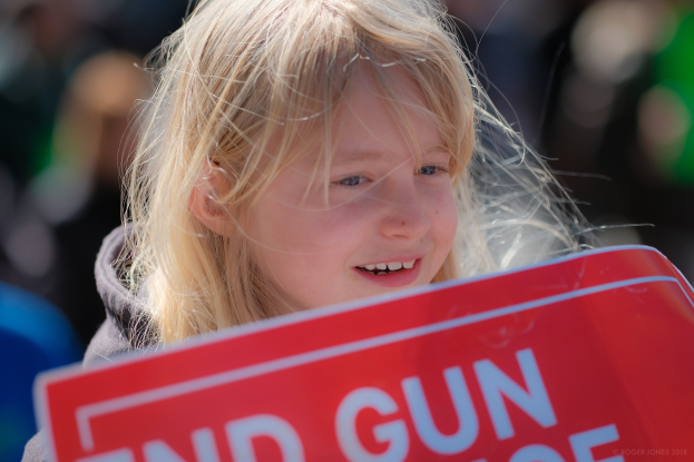 Ein junges Mädchen mit strahlendem Lächeln hält ein rotes Schild hoch, auf dem «End Gun Violence» steht, vor einem unscharfen Hintergrund.