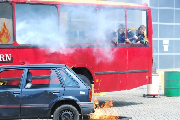 Roter Doppeldeckerbus mit Rauch und drei sichtbaren Passagieren, neben einem Auto geparkt, vor einem Glasfenster-Gebäude mit einem Fass auf der rechten Seite.