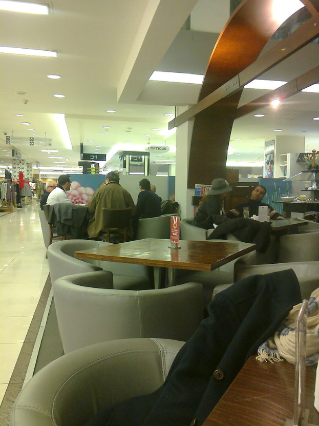 Menschen auf Stühlen in einem Einkaufszentrum, mit Kleidung auf einem Tisch, Stores auf der linken Seite und Lampen an der Decke.