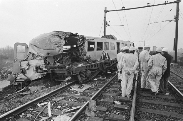 Ein Schwarz-Weiß-Bild einer Zugkollision mit Menschen in Helmen, die darum herumstehen, Strommasten, Bäumen, Gebäuden und einem klaren Himmel im Hintergrund.