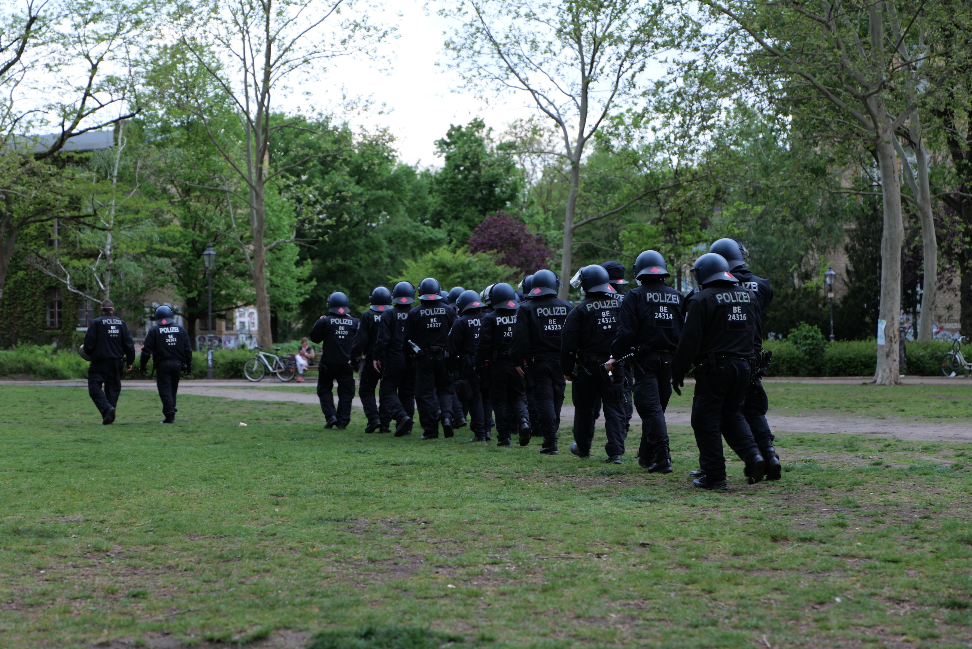 Group of police officers in black uniforms and helmets walking across a green field with bicycles, light poles, plants, trees, buildings, and a clear blue sky in the background.