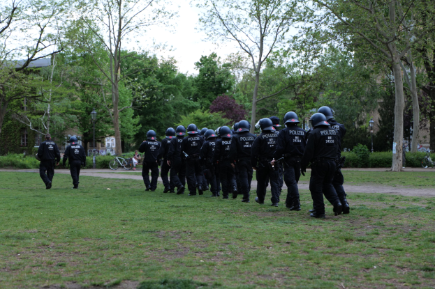 Group of police officers in black uniforms and helmets walking across a green field with bicycles, light poles, plants, trees, buildings, and a clear blue sky in the background.