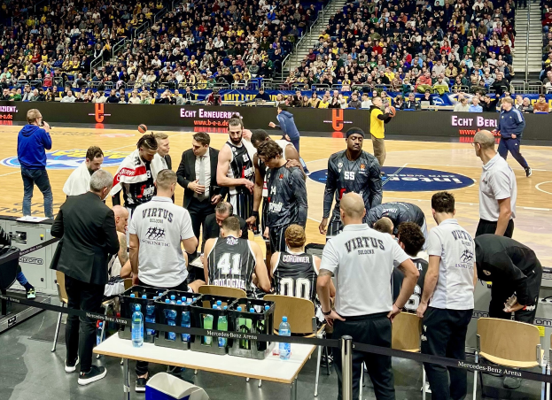 Eine Gruppe von Männern auf einem Basketballfeld mit einer Menge drumherum, ein Fotograf mit Equipment auf einem Tisch und Hintergrundtafeln mit Teamnamen Virtus Berlin und Echt Berlin.