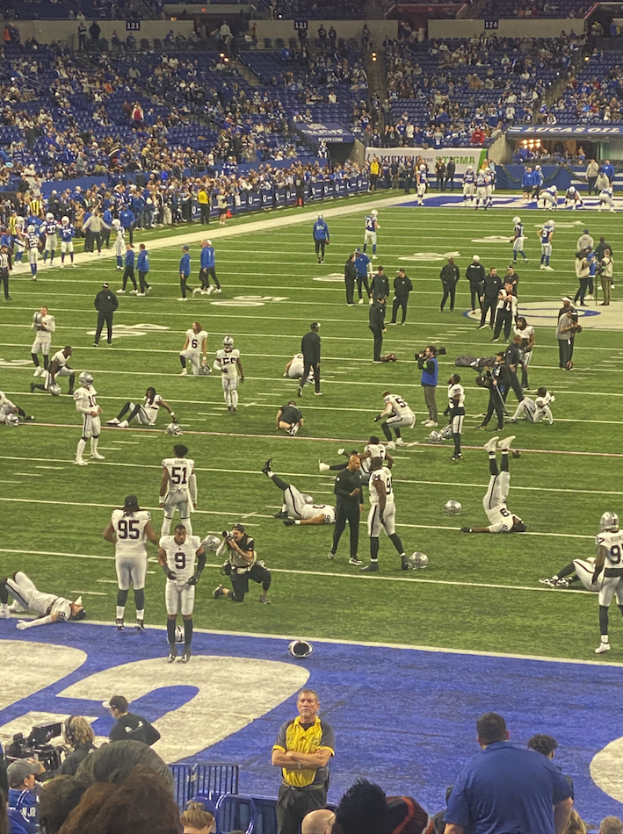 Ein Fußballspiel wird in einem Stadion mit Zuschauern und einigen auf Stühlen sitzenden Personen sowie einer Person mit einer Kamera gespielt, was auf eine Live-Übertragung hinweist.