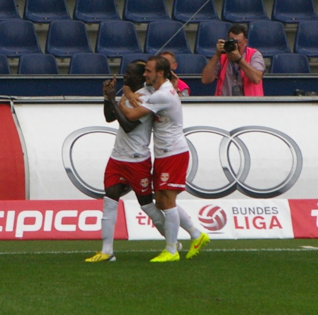 Zwei Fußballspieler feiern ein Tor auf einem Feld mit einer Gruppe von Menschen drumherum, eine Person hält eine Kamera, eine Fahne mit Text, Stühle und eine Wand im Hintergrund.