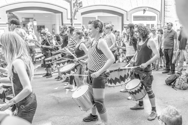 Eine Gruppe von Menschen, die musikalische Instrumente im Freien spielen, vor einer geschlossenen Glastür mit Licht im Hintergrund.