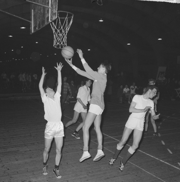 Schwarzes und weißes Foto von jungen Männern, die Basketball in einer Turnhalle mit einem Netz und Zuschauern im Hintergrund spielen.