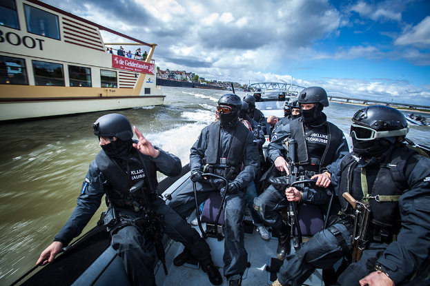 Eine Gruppe von Polizisten in schwarzen Uniformen und Helmen, einige bewaffnet, fahren auf einem Boot auf dem Wasser mit einer Brücke, Bäumen, Gebäuden und einem klaren blauen Himmel im Hintergrund.