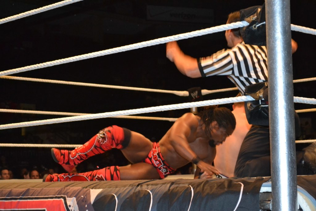 Zwei Männer kämpfen in einem Ring, einer trägt ein rotes Hemd und Schuhe, der andere Shorts, daneben ein Schiedsrichter und eine Zuschauermenge im Hintergrund unter einem dunklen Himmel.