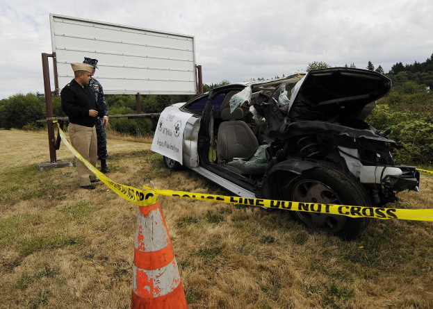 Ein Polizeibeamter in Uniform steht neben einem schwer beschädigten Auto an der Straßenseite, mit eingedrückter Motorhaube und zersplitterten Fenstern, unter einem bewölkten Himmel.
