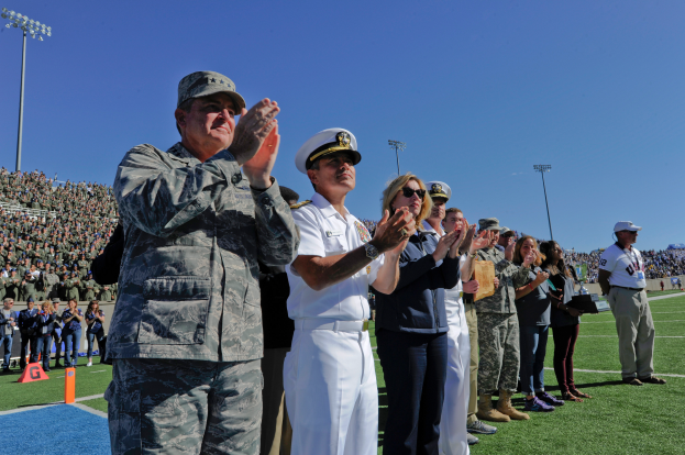 Eine Gruppe von Menschen auf einem Fußballfeld klatschen, mit Zuschauern auf den Tribünen, Flutlichtmasten und einem klaren blauen Himmel im Hintergrund.