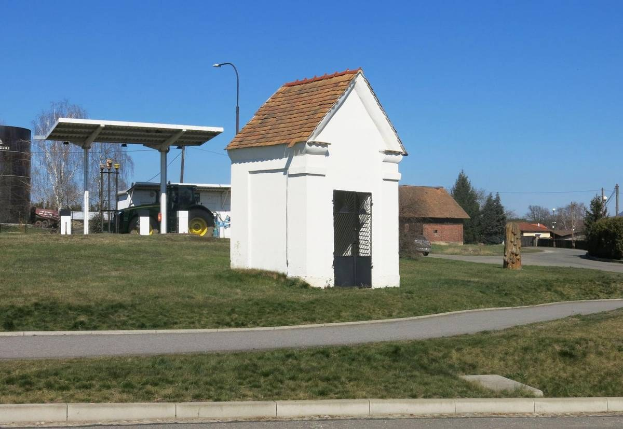 Kleines weißes Gebäude mit rotem Dach, erste Tankstelle in Deutschland, auf einer grasbewachsenen Straße mit Bäumen, Pfählen und Häusern unter einem klaren blauen Himmel.
