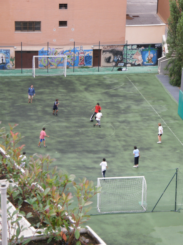 Kinder spielen Fuball auf einem Feld mit Toren, ein Graffiti-beschmiertes Geb"ude im Hintergrund, Pflanzen links und B"ume rechts.