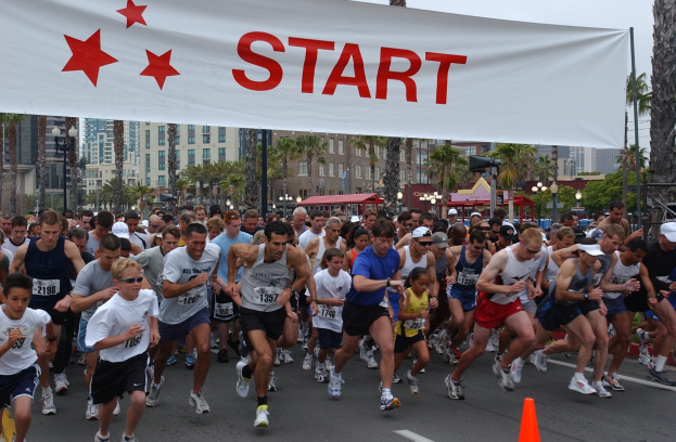 Eine Gruppe von Menschen, die bei einem Marathon laufen, mit einem Verkehrskegel im Vordergrund und einer Fahne im Hintergrund, umgeben von Bäumen, Laternenmasten, Gebäuden und einem klaren blauen Himmel.