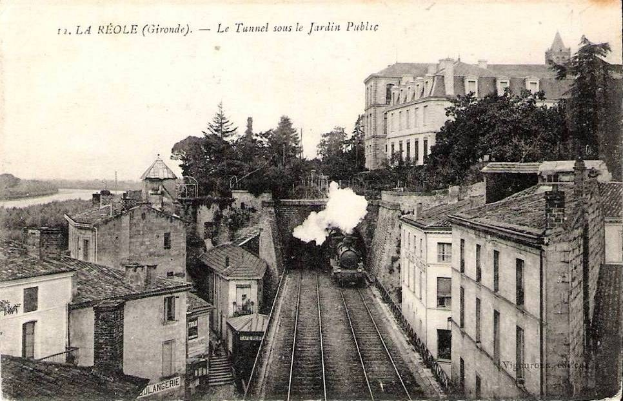 Black and white photograph of a train on tracks near buildings, trees, and water with text "la réole gironde - le tunnel sous le jardin public" at the top.