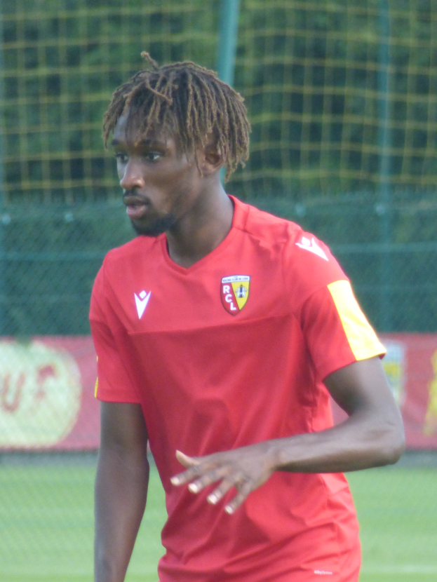 Ein Mann mit Dreadlocks steht auf einem Fussballfeld und trägt ein rotes T-Shirt und Shorts, im Hintergrund gibt es ein Netz und eine Fahne mit der Aufschrift "RCCL - Ghanaian Premier League".
