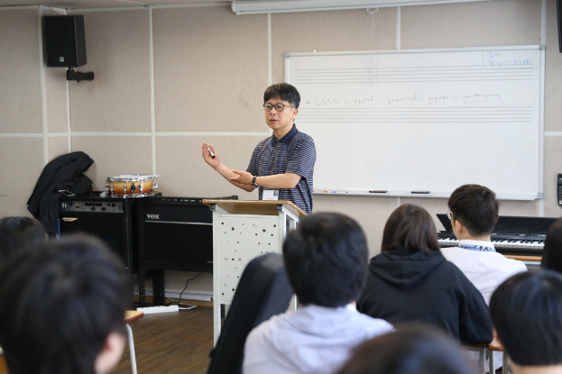 Ein Mann steht an einem Rednerpult und spricht zu einem Publikum, mit einer Whiteboard und einem wandmontierten Lautsprecher dahinter.