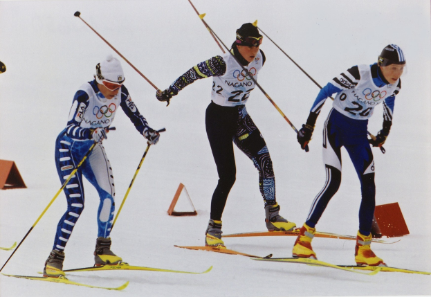 Drei Skifahrer in buntfarbigen Anzügen und Helmen fahren eine schneebedeckte Piste hinunter, jeder hält Ski-Stöcke.