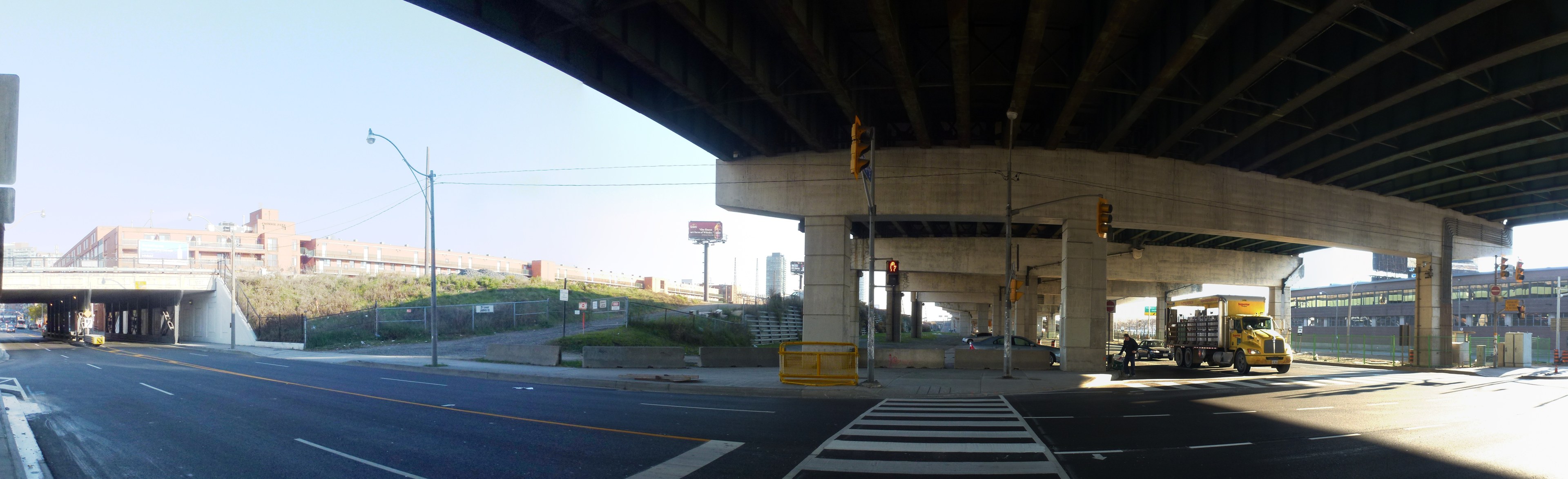 A truck drives under a bridge on a city street with buildings, street furniture, vehicles, greenery, and a clear sky.