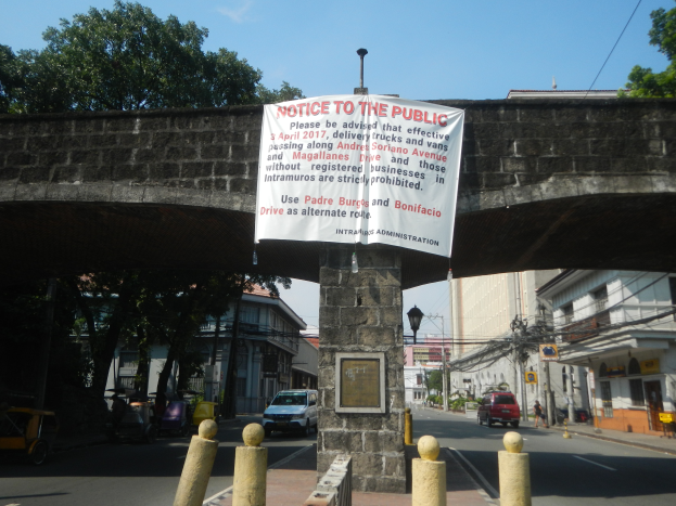 Eine Brücke mit einem "Hinweis"-Schild, Fahrzeuge auf der Straße darunter und Gebäude, Bäume, Pfähle, Drähte und einen klaren blauen Himmel im Hintergrund.