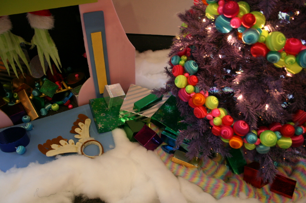 Eine Indoor-Szene mit einer Wand im Hintergrund, einem Baumwoll- und Wolltuch am Boden, einem geschm├╝ckten Weihnachtsbaum rechts, Geschenkboxen und Deko-Artikeln in der Mitte und einer H├╝tte mit Deko und Baumwolle oben links.