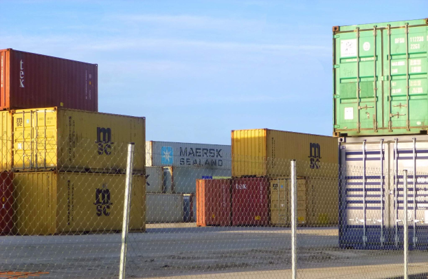 Lagercontainer in Reihen mit Maschendrahtzaun im Vordergrund, Straße unten und bewölkter Himmel im Hintergrund.