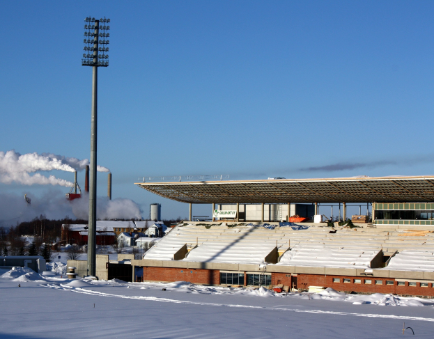 Ein schneebedecktes Feld mit einem Stadion und umliegenden Gebäuden, Bäumen, Mästen, Lichtern und Rauch, unter einem sichtbaren Himmel.