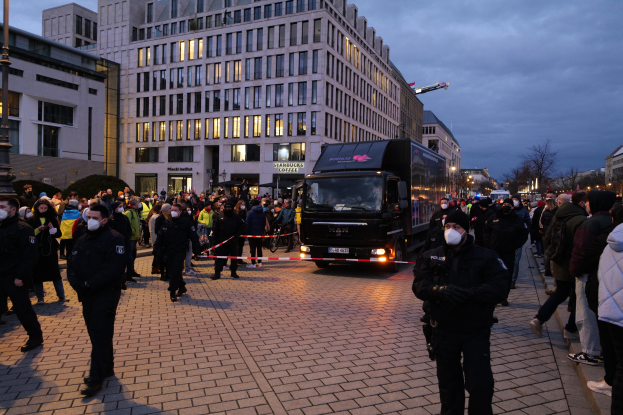 Gruppe von Menschen vor einem Lastwagen auf einer Straße mit Gebäuden, Bäumen und Laternen; einige tragen Mützen und Masken, mit einem Band an einem Pfahl im Vordergrund.