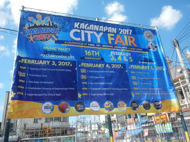 Plakat mit 'Kaganapan City Fair 2017'-Text, umgeben von Pfählen, Drähten, Gebäuden, Bäumen, einem Zaun und einem klaren blauen Himmel.
