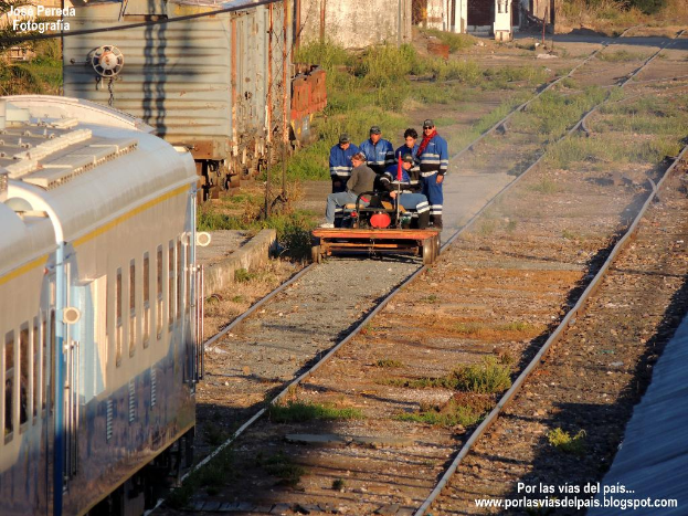 Eine Gruppe von Menschen auf einem Fahrzeug in der Nähe von Eisenbahnschienen, ein Zug, ein Schuppen, Eisenstangen, Pflanzen und eine Flagge.