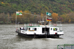 Eine Fähre fährt einen Fluss hinunter, mit einem Geländer auf der rechten Seite und einem Hügel mit Bäumen im Hintergrund; das Boot ist mit Fahnen, Stangen und anderen Gegenständen geschmückt und ein paar Menschen sind an Bord.