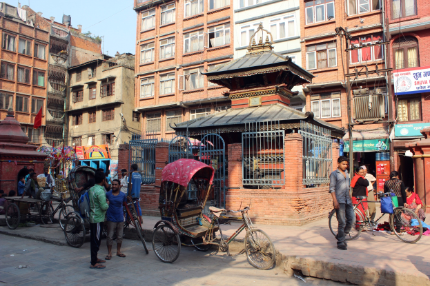 A bustling street in Kathmandu, Nepal, with pedestrians, rickshaws, bicycles, and vehicles alongside tall buildings under a clear blue sky.