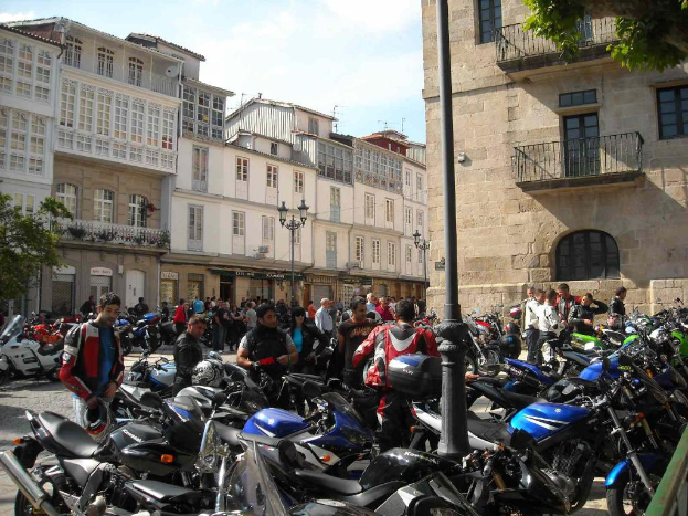 Ein cremefarbenes Gebäude mit zahlreichen Fenstern, neben einem Backsteingebäude mit einer Metallrahmen-Balkon, mit mehreren geparkten Fahrrädern und Menschen, die vorne diskutieren.