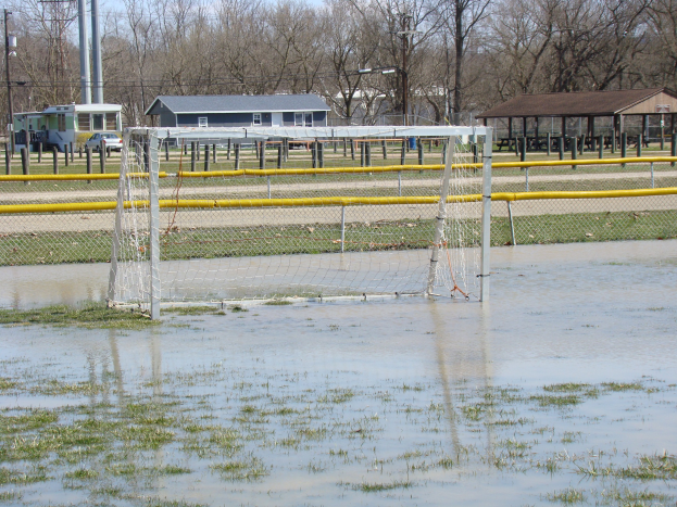 Ein Fußballtor steht in der Mitte eines überfluteten Feldes, mit Hütten, Pfählen, Bäumen und Fahrzeugen im Hintergrund unter einem klaren blauen Himmel.