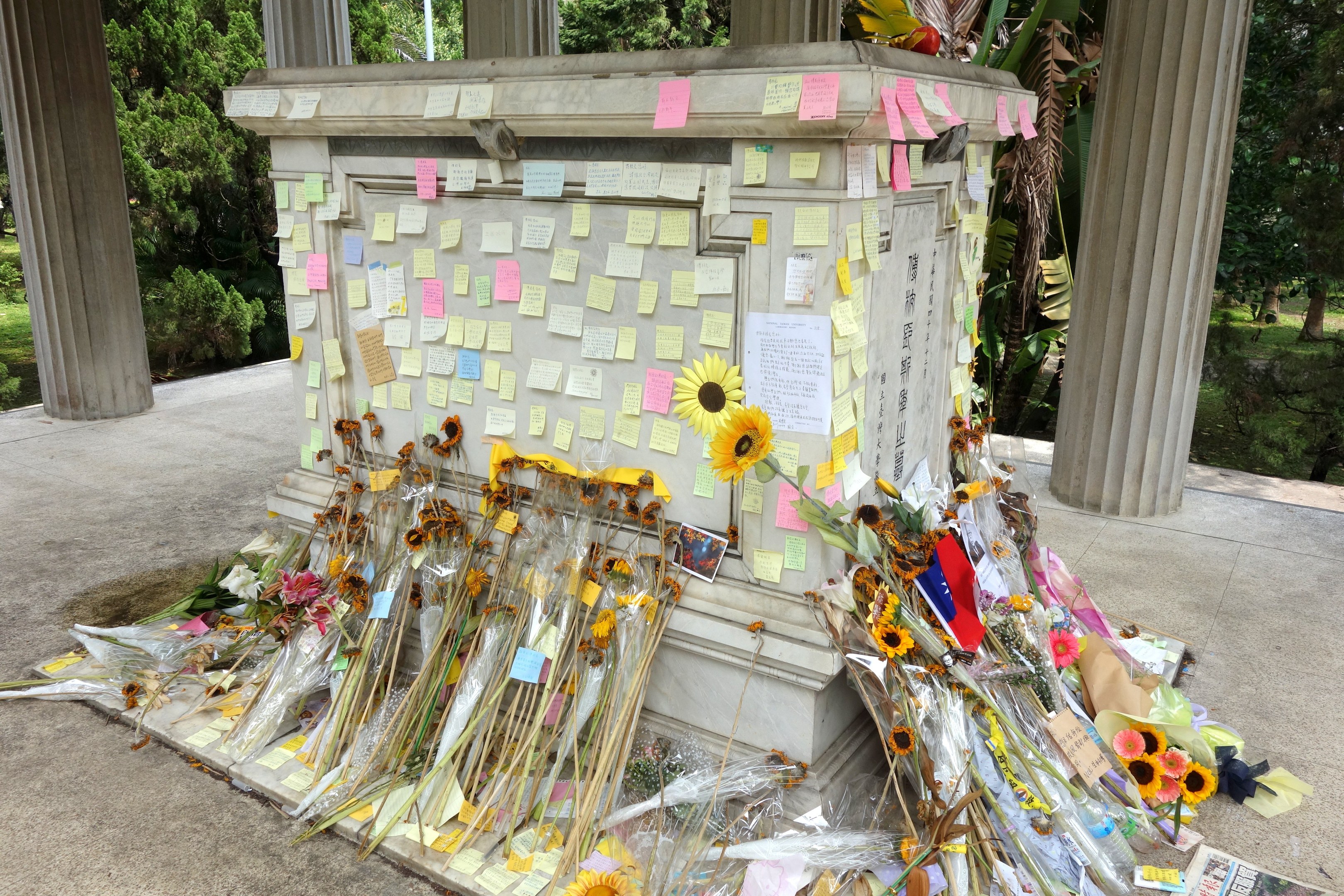 Gedenken mit einem zentralen Pfeiler, der mit Post-it-Notizen und Blumen geschmückt ist und von Bäumen, Pflanzen und Gras umgeben ist.