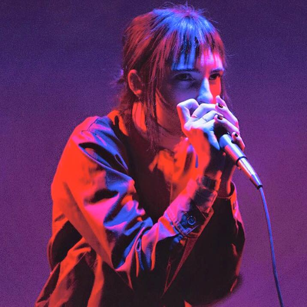 Eine Frau in einem schwarzen Kleid singt in ein Mikrofon auf einer Bühne, die von lila und blauen Lichtern beleuchtet wird, mit geschlossenen Augen und leicht geöffneten Lippen.