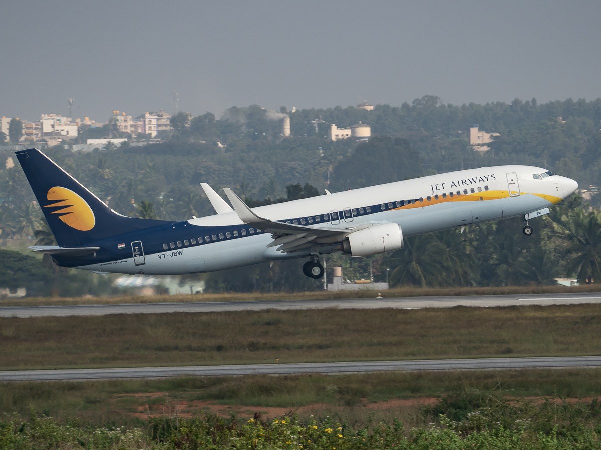 A Jet Airways plane taking off from an airport runway with green grass, trees, buildings, and a clear blue sky in the background.