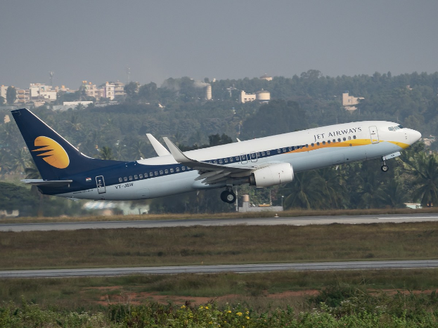 A Jet Airways plane taking off from an airport runway with green grass, trees, buildings, and a clear blue sky in the background.