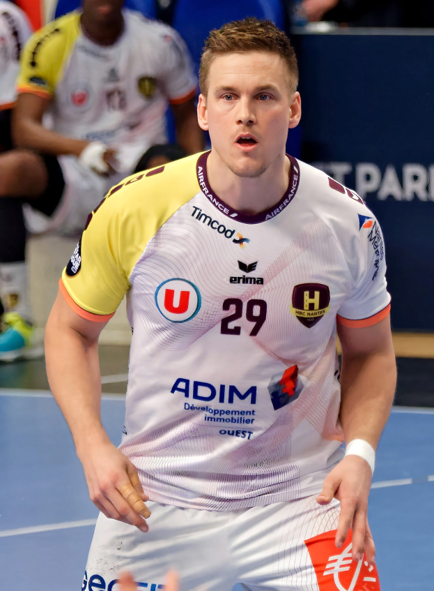 Ein Mann in weißem Hemd und gelben Shorts spielt Handball, hält ein Objekt und hat einen entschlossenen Gesichtsausdruck, während im Hintergrund Menschen auf dem Boden sitzen und eine Tafel die Aufschrift 'Ligue 1 - Futsal - Olympique de Marseille' trägt.