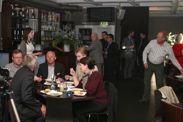 Eine Bar-Inneneinrichtung mit Flaschen in einem Schrank, einer Schautafel, einem Fernseher, einem Vorhang, einer Vase, stehenden Menschen und anderen an einem Tisch sitzend mit Essen und Getränken.