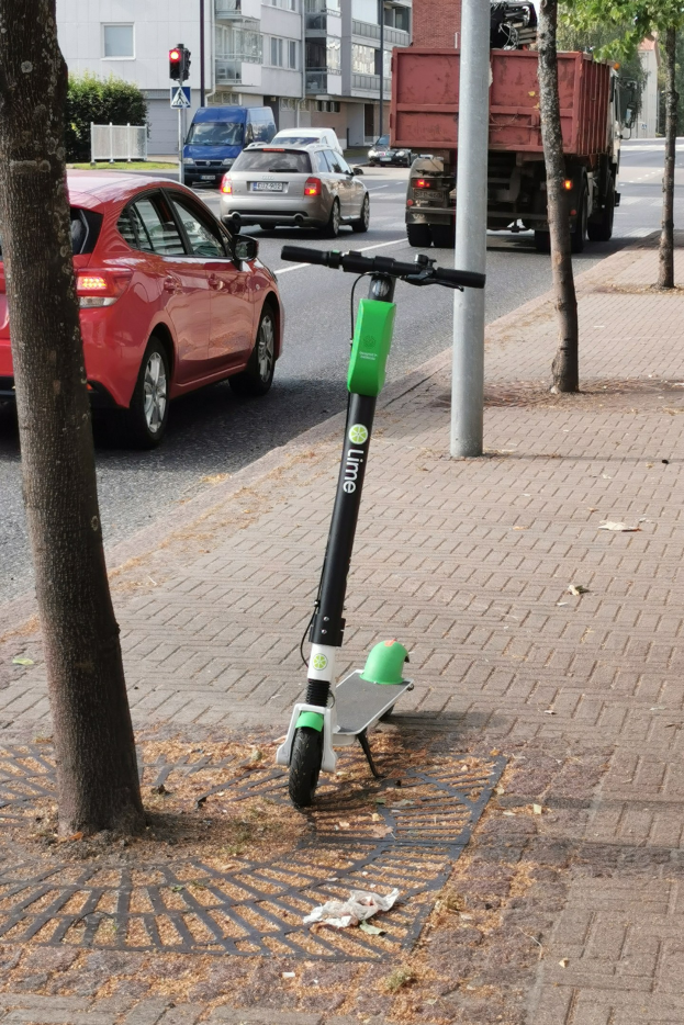 Elektroroller auf einem Gehweg neben einem Baum geparkt, mit Fahrzeugen auf der Straße und Gebäuden im Hintergrund.