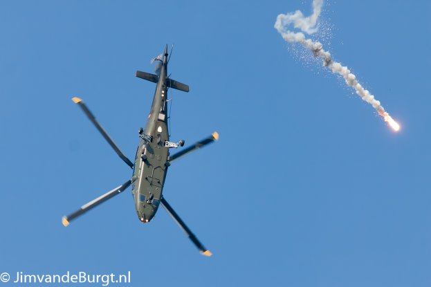 Ein militärisches Hubschrauber in der Luft, das eine Rakete hält, die Rauch und Feuer abgibt, mit Text in der unteren linken Ecke.