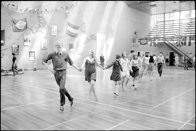 Schwarz-weißes Bild von Menschen, die in einem Turnsaal tanzen, mit Fotoeinrahmen und Fahnen an der Wand, Treppe auf der rechten Seite sichtbar.