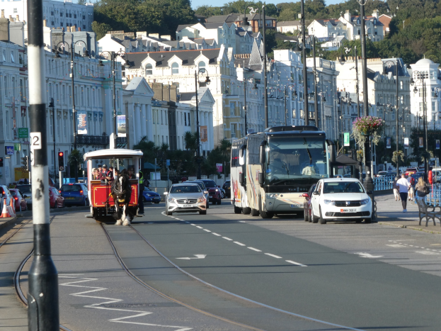 Eine belebte Stadtstraße mit hohen Gebäuden, Fahrzeugen, Fußgängern, Ampeln, Laternenmasten, Texttafeln, Bäumen und einem klaren blauen Himmel.