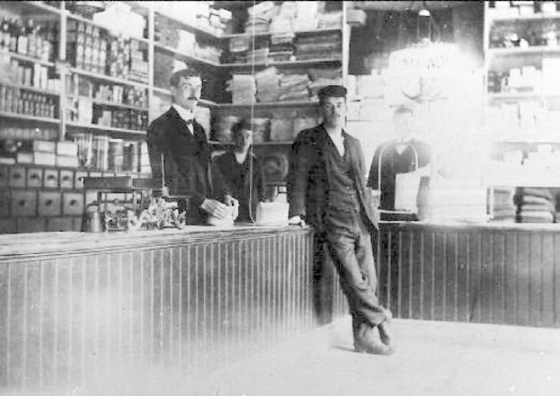Schwarz-weißes Foto von drei Männern an einer Ladentheke während eines scheinbaren Raubs, mit Büchern und Gegenständen im Hintergrund.