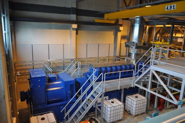 Ein Fabrikinnenraum mit Trägern, einer Treppe mit Geländer, Containern, Lampen, Rohren und Wänden.